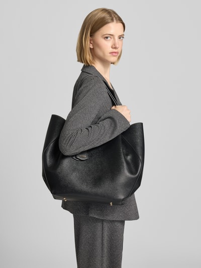 Frauenmodel trägt Tote Bag in Leder-Optik Modell 'Amorette' von GUESS, schwarz, Logo-Detail, Nahaufnahme Oberkörper. Hauptfach mit Magnetverschluss, schmale Schulterriemen, 44x37x11 cm, Riemen 56 cm.