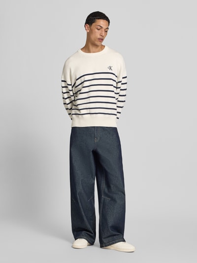 Männermodel trägt Regular Fit Strickpullover aus reiner Baumwolle von Calvin Klein Jeans, gestreifte look, gestikt logo, lange mouw, ronde hals, rompzoom geribd, kleur gebroken wit met donkerblauwe strepen, gebreide textuur, heren gebreide pullover, materiaalverwerking gebreid, puur katoen, ganzkörperansicht von vorne.