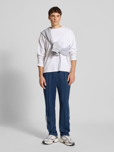 Männermodel trägt Trackpants mit elastischem Bund Modell 'LATUCCI' von Ellesse in Dunkelblau mit weißen Galonstreifen, lockerer Passform, Ganzkörperansicht von vorne.