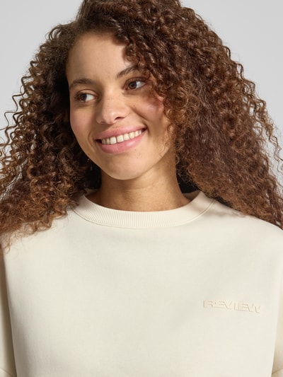 Modelka nosi Basic Sweater mit Rundhalsausschnitt und Bündchen am Ärmel od REVIEW, kremowy, gładki, logo, oversize, zbliżenie na górę ciała.
