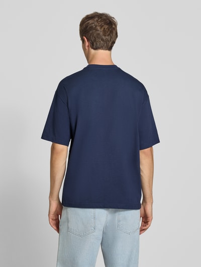 Männermodel trägt Relaxed Fit T-Shirt aus reiner Baumwolle von Champion in Dunkelblau, glatte Textur, Rückenansicht, Ganzkörperansicht Rückansicht. Rückenlänge bei Größe M 70 cm, Logo-Print, Motiv-Stitching, Rundhalsausschnitt.