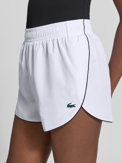 Frauenmodel trägt Regular Fit Sportshorts mit elastischem Bund von Lacoste in Weiß mit schwarzer Paspel, glatte Textur, Nahaufnahme Oberschenkel, Damen Sportshorts von Lacoste Sport.