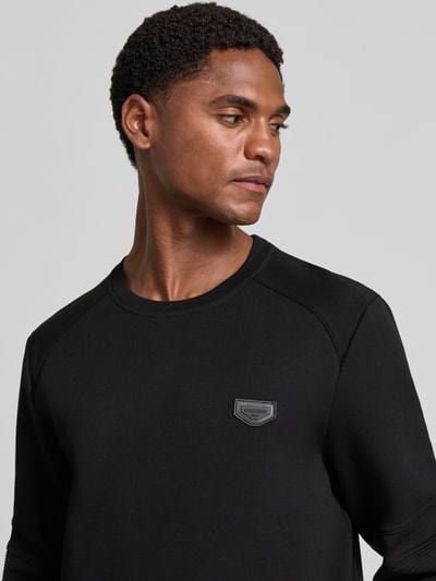 Mannenmodel draagt zwart sweatshirt met labelpatch Model 'NEUTER' van Antony Morato, geribde ronde hals, detailopname bovenlichaam. Heren, getailleerd, mouwen met versteviging, elastische boorden, ruglengte maat M 66 cm, mouwlengte maat M 58 cm.