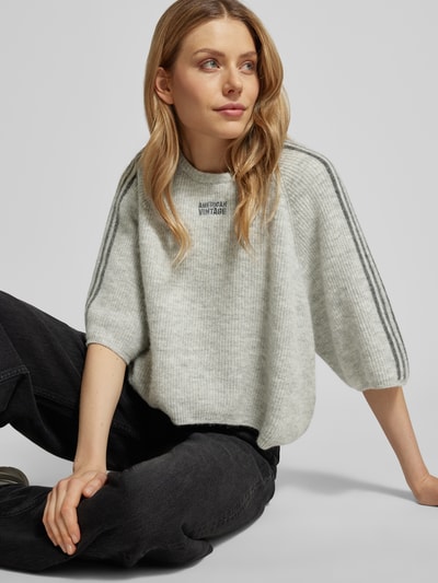 Vrouwenmodel draagt Cropped Strickpullover met geribde boorden Modell 'VITOW' van American Vintage in wol-alpaca-mix, los vallende fit, ronde hals, label-stitching, lichtgrijs met donkere strepen, materiaal met stretch, vooraanzicht zittend, context: damespullover ruglengte 53 cm, mouwlengte 36 cm