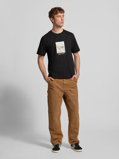 Männermodel trägt T-Shirt mit Rundhalsausschnitt Modell 'JORNORREBRO' von Jack & Jones aus reiner Baumwolle, gerade geschnitten, schwarze Farbe mit Frontprint, kombiniert mit braunen Hosen, Ganzkörperansicht von vorne.