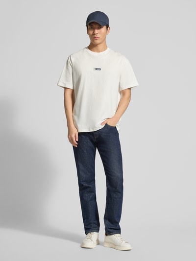 Mężczyznamodel nosi T-Shirt mit Logo-Print Modell 'Chase' od Les Deux z czystej bawełny, krój regular fit, biały kolor, nadruk logo, detal z metką, długość tyłu w rozmiarze M: 72 cm, całopostaciowy widok z przodu.