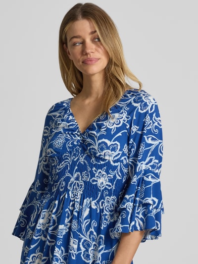 Frauenmodel trägt Minikleid aus reiner Viskose mit Smok-Detail von Apricot, floraler Allover-Print in Blau und Weiß, Rüschenbesatz, 3/4-Arm, V-Ausschnitt, taillierte Passform, Nahaufnahme Oberkörper.