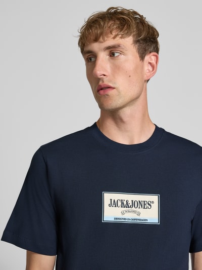 Męski model nosi t-shirt z nadrukiem z logo Model 'FREDERIKSBERG' od JACK & JONES, bawełna, prosty krój, okrągły dekolt, długość tyłu w rozmiarze M: 70 cm, zbliżenie na górną część ciała.