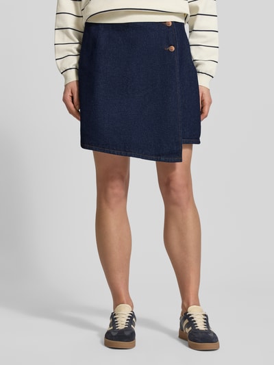Frauenmodel trägt Regular Fit Minirock in Wickel-Optik Modell 'Lisana' von YAS, reine Baumwolle, Denim-Optik, asymmetrisches Design, 2-Knopf-Leiste, Ganzkörperansicht von vorne.