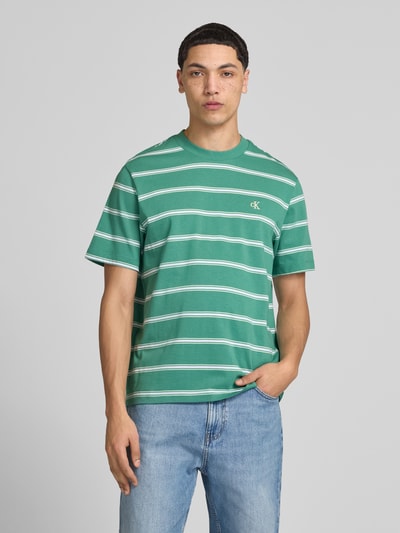Männermodel trägt T-Shirt mit Logo-Stitching von Calvin Klein Jeans in Grün mit weißen Streifen, Reine Baumwolle, Gerader-Schnitt, Rundhalsausschnitt, Rückenlänge 69 cm bei Größe M, Ganzkörperansicht von vorne.