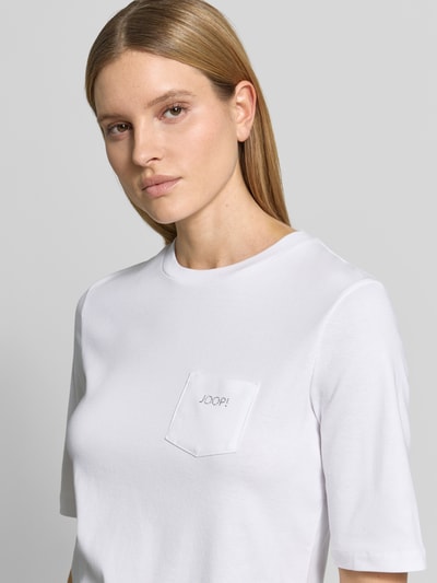 Frauenmodel trägt Regular Fit T-Shirt mit Brusttasche Modell 'Tonja' von JOOP! in Weiß, glatte Baumwolle, Strasssteinbesatz, Nahaufnahme Oberkörper.