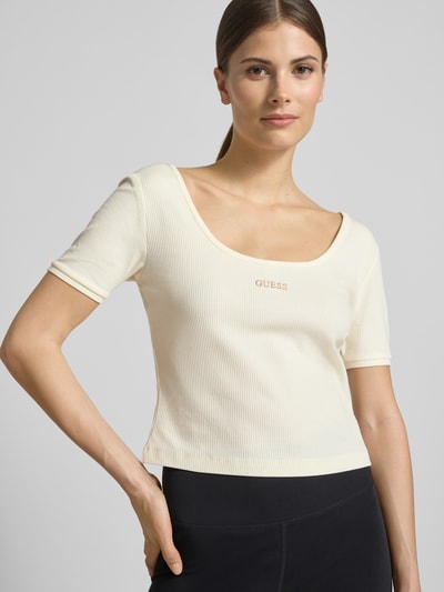 Frauenmodel trägt Cropped T-Shirt aus Lyocell-Mix Modell 'Circe' von Guess Activewear in Creme mit Logo-Stitching, Nahaufnahme Oberkörper.