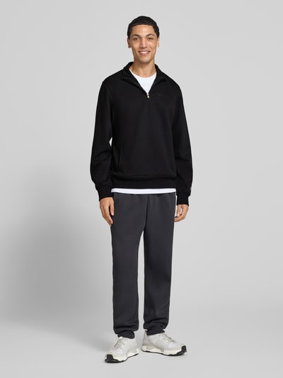 Männermodel trägt Sweatshirt mit Label-Details von Champion in Schwarz, Baumwollmischung, Troyer-Kragen, seitliche Taschen, Ganzkörperansicht von vorne.