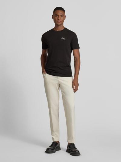 Männermodel trägt T-Shirt mit Label-Print von EA7 Emporio Armani in Schwarz, Slim Fit, glatte Struktur, Ganzkörperansicht von vorne.