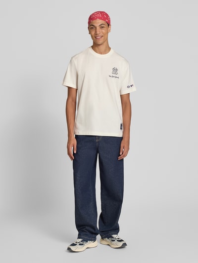 Männermodel trägt Relaxed Fit T-Shirt aus reiner Baumwolle von Champion mit Statement-Print, hellem Stoff, kombiniert mit Jeans, Ganzkörperansicht von vorne.