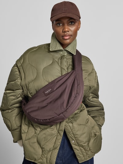 Frauenmodel trägt Crossbody Bag aus Teddyfell mit Logo-Detail von Studio Noos, olivgrüne Steppjacke, braune Cap, Nahaufnahme Oberkörper.