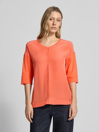 Modelka kobieca nosi damski t-shirt 'T-Shirt mit Label-Detail' od Marc Cain z mieszanki bawełny i elastanu, prosty krój, dekolt w serek, ozdobne szwy, rękaw o dł. 3/4, detal z logo, długość tyłu w rozmiarze 36: 60 cm, kolor koralowy, gładka faktura, widok całej sylwetki z przodu.