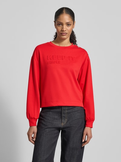 Frauenmodel trägt Sweatshirt mit Viskose-Anteil von monari in Rot, Rundhals mit Nieten, Statement-Prägung, lockere Passform, Nahaufnahme Oberkörper.