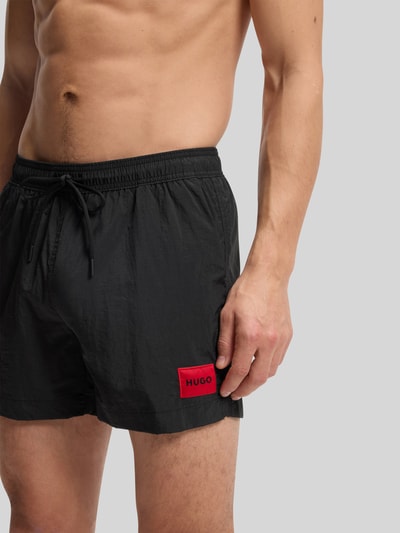 Männermodel trägt schwarze Badeshorts mit Label Patch Modell 'DOMINICA' von HUGO, unifarben, elastischer Bund, Nahaufnahme Oberkörper.