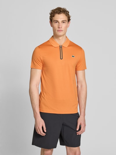 Männermodel trägt Slim Fit Poloshirt aus schnelltrocknendem Material von Lacoste Sport in Orange mit Logo-Patch, Ganzkörperansicht von vorne.