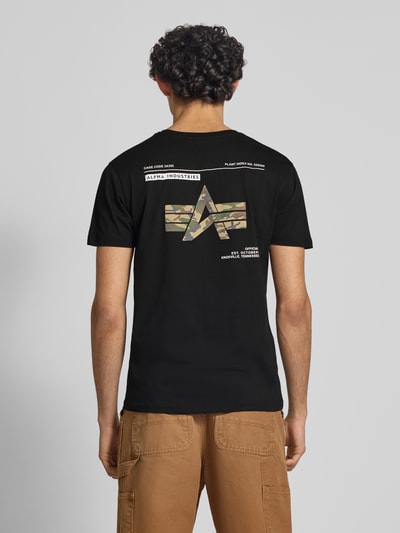 Männermodel trägt T-Shirt mit Label-Patch Modell 'Camo' von Alpha Industries aus schwarzer, gewachster Baumwolle mit Camouflage-Print; Ganzkörperansicht Rückansicht