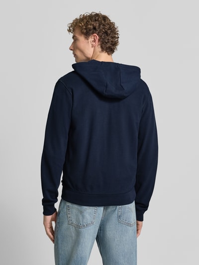 Męski model nosi granatową bluzę rozpinaną Relaxed Fit Sweatjacke aus reiner Baumwolle od s.Oliver RED LABEL, widok całej sylwetki z tyłu.