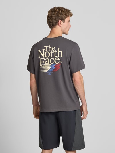 Männermodel trägt Relaxed Fit T-Shirt mit Logo-Print von The North Face in Dunkelgrau mit mehrfarbigem Print, reine Baumwolle, Ganzkörperansicht Rückansicht.