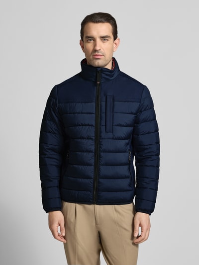 Męski model nosi Regular Fit Steppjacke mit seitlichen Stretcheinsätzen od Tom Tailor, granatowa, steppowana, krój regular fit, stójka, zamek, logo, elastyczne wykończenia, wodoodporna, kieszenie, widok całej sylwetki z przodu.