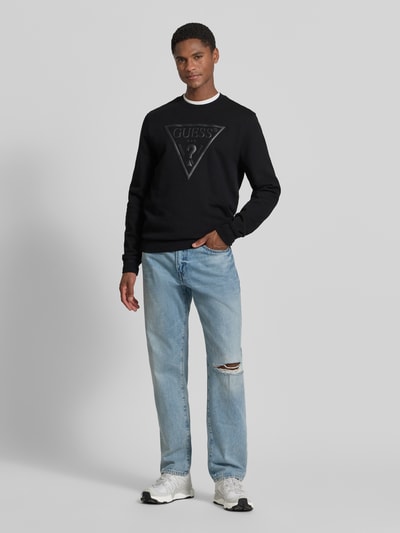 Männermodel trägt Regular Fit Sweatshirt mit Logo-Detail Modell 'Audley' von Guess aus Baumwoll-Mix, schwarze Farbe, Ganzkörperansicht von vorne. Herren Sweatshirt mit Rundhalsausschnitt und elastischen Abschlüssen, Rückenlänge 69 cm, Ärmellänge 69 cm, dezent strukturierte Oberfläche.