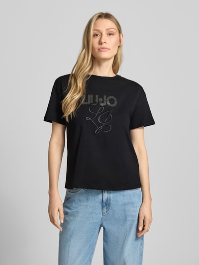 Kobiece model prezentuje T-Shirt mit Label-Stitching und Ziersteinbesatz od Liu Jo White z bawełny, krój regular fit, okrągły dekolt, długość tyłu 56 cm, całkowity widok z przodu.