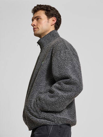 Männermodel trägt Regular Fit Jacke mit Logo-Patch von American Vintage, Kunstfell in Grau, Eingrifftaschen, Stehkragen, Nahaufnahme Oberkörper.