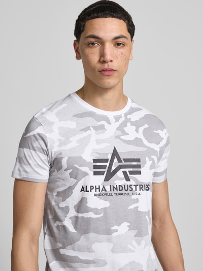 Męski model nosi T-Shirt mit Label-Print od Alpha Industries w kolorach szaro-białych, bawełna, krój regular fit, nadruk logo, zbliżenie na górną część ciała.