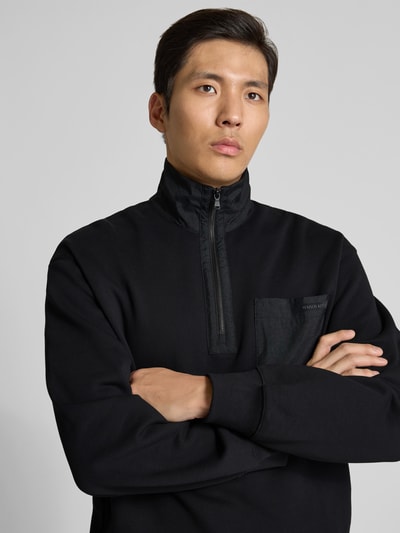Mannenmodel draagt Troyer aus reiner Baumwolle van MAISON KITSUNÉ in zwart met labeldetail, borstzak, regular fit, puur katoen, nahaufnahme bovenlichaam.