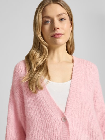 Frauenmodel trägt Cardigan mit V-Ausschnitt Modell 'EAST' von American Vintage in Hellrosa, grobe Strickstruktur, Nahaufnahme Oberkörper. Relaxed Fit, Knopfleiste, Rückenlänge 62 cm, Ärmellänge 48 cm bei XS/S.