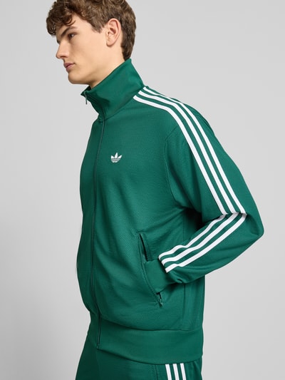 Mężczyzna model nosi Trainingsjacke mit Stehkragen od adidas Originals, kolor zielony, pasek biały, krój regular fit, zbliżenie na górną część ciała, widok z przodu.