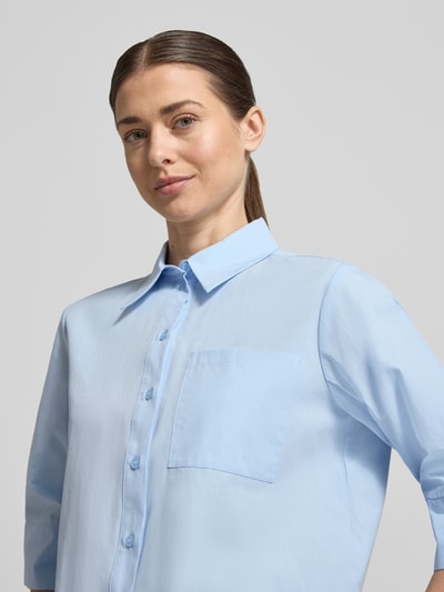 Modelka kobieca nosi Boxy Fit Bluse mit Brusttasche Modell 'Zarba Bold' od Someday, jasnoniebieska bawełna, kieszeń na piersi, kołnierzyk, krótki rękaw, długość tyłu 54 cm w rozm. 36, długość rękawa 39 cm w rozm. 36, zbliżenie na górną część ciała