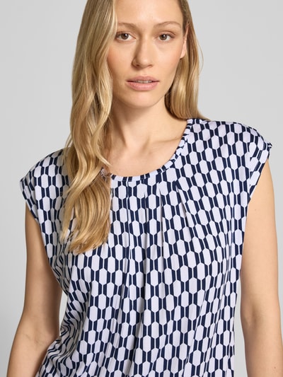 Vrouwenmodel draagt Blusenshirt mit Rundhalsausschnitt van ZABAIONE in mix van viscose en elastaan met recht model, korte mouwen, ruglengte maat S 58 cm, geometrisch blauw-wit patroon, textuur soepel, Nahaufnahme Oberkörper.