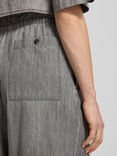 Frauenmodel trägt Wide Fit Stoffhose mit Rüschenbesatz Modell 'Dwindle' von Drykorn aus reiner bawełna, wpuszczane kieszenie, elastyczny pas z tunelem, obszycie falbanami, szerokość w rozmiarze 27/34: 37 cm, długość wewnętrzna nogawki: 82 cm, szary materiał, tekstura jeansu, detal tyłu spodni, Nahaufnahme okolicy kieszeni z tyłu.