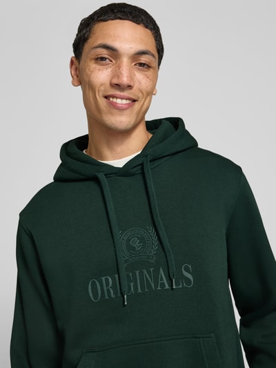 Männermodel trägt Hoodie aus Baumwoll-Mix Modell 'FREDERIKS' von JACK & JONES in Dunkelgrün mit Label-Print, Nahaufnahme Oberkörper.
