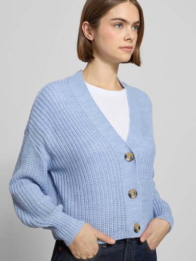 Frauenmodel trägt Regular Fit Strickjacke mit einreihiger Knopfleiste Modell 'CAROLSPRING' von Only in Unifarben Strickoptik, Cropped-Länge, Label-Stitching-Applikation, tiefer V-Ausschnitt, Langarm, gerippter Ärmel- und Rumpfabschluss, Materialverarbeitung: Strick, Nahaufnahme Oberkörper.