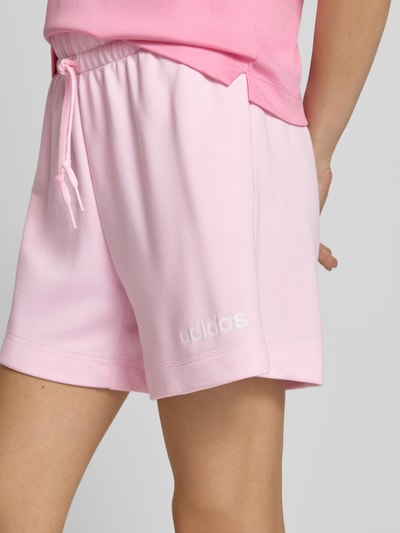 Vrouwenmodel draagt Shorts met elastische tailleband en trekkoord van adidas Sportswear, lichtroze, katoenmix, regular fit, logo, detailopname heup.