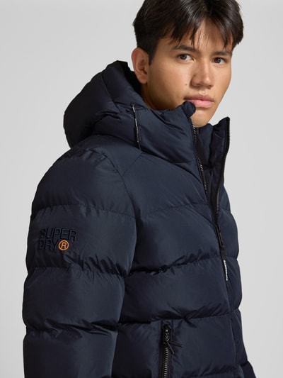 Männermodel trägt Steppjacke mit Label-Stitching und Kapuze von Superdry in Dunkelblau, glatte Steppstruktur, Nahaufnahme Oberkörper. Herren Steppjacke, gerader Schnitt, Kapuze mit Kordelzug, Reißverschluss, seitliche Eingrifftaschen, elastische Ärmelabschlüsse, Rückenlänge 67 cm, Schulterbreite 48 cm, Brustweite 58 cm, Ärmellänge 72 cm.