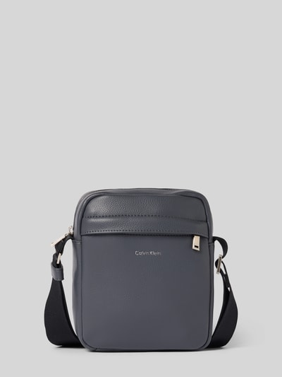 Produktfoto, graue Handtasche mit Label-Detail Modell 'RAISED REPORTER' von CK Calvin Klein, Hauptfach mit Reißverschluss, Schulterriemen, Maße 17x20x7 cm, Länge Schulterriemen 68 cm