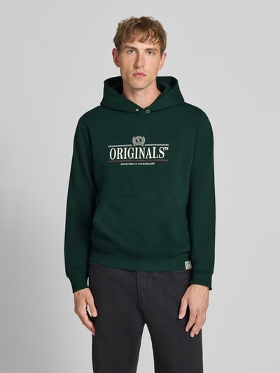 Männermodel trägt Hoodie mit Label-Print Modell 'FREDERIKSBERG' von JACK & JONES in Dunkelgrün, Baumwoll-Mix, Ganzkörperansicht von vorne. Kontext: Lockere Passform, Kapuze, Label-Print, Kängurutasche, gerippte Abschlüsse, Rückenlänge 65 cm, Ärmellänge 67 cm.
