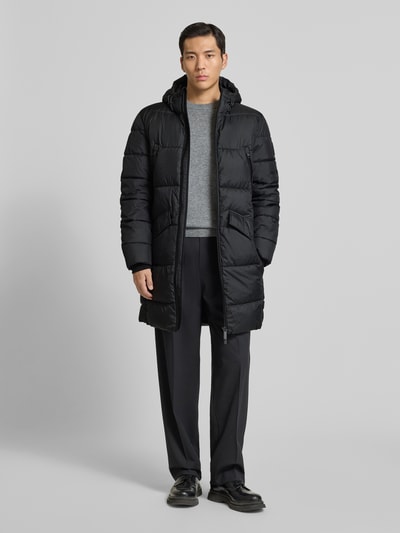 Męski model ma na sobie Regular Fit Steppmantel mit Kapuze von Tom Tailor, krój regular fit, czarny, długie rękawy, kieszenie na zamek, stehkraag z kapturem, łatka z logo, przód zapinany na dwukierunkowy zamek błyskawiczny, widok całej sylwetki z przodu.