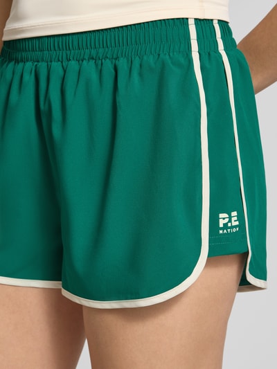Frauenmodel trägt Relaxed Fit Shorts mit integriertem Slip von P.E Nation, grün mit weißem Logo-Detail und elastischem Bund, Nahaufnahme Oberschenkel. Shorts für Damen mit Bundweite 37 cm und Innenbeinlänge 6 cm in Größe S.