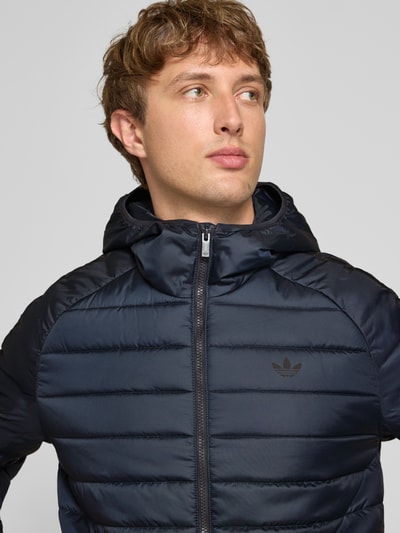 Männermodel trägt Regular Fit Steppjacke mit Reißverschluss von adidas Originals in Dunkelblau, glatte und gesteppte Struktur, Nahaufnahme Oberkörper.
