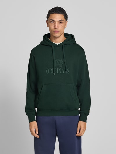Männermodel trägt Hoodie aus Baumwoll-Mix Modell 'FREDERIKS' von JACK & JONES in Dunkelgrün mit Label-Print, Ganzkörperansicht von vorne. Locker geschnitten, gerippte Abschlüsse, Kapuze mit Tunnelzug, Kängurutasche, Rückenlänge 67 cm, Ärmellänge 65 cm.