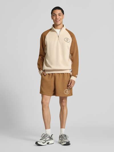 Männermodel trägt Regular Fit Sweatshorts aus Baumwoll-Mix von Nike in Braun mit Logo, kombiniert mit Sweatshirt, Ganzkörperansicht von vorne.