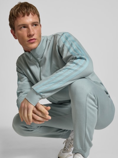 Männermodel trägt Sweatanzug mit Label-Stitchings von adidas Sportswear aus Baumwoll-Mix in hellblau, Nahaufnahme Oberkörper. Regular-Fit, Sweatjacke mit Stehkragen, Reißverschluss, Hose mit Label-Stitching, Bundweite bei Größe M 73 cm, Rückenlänge 72 cm, Schulterbreite 50 cm, Ärmellänge 65 cm, Innenbeinlänge 72 cm.